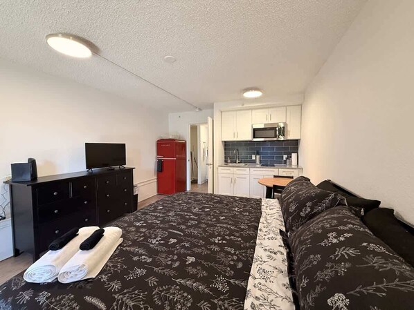 Un dormitorio con cama, televisión, cómoda, refrigerador rojo, área de cocina con armarios y microondas, y baño con inodoro y lavamanos.