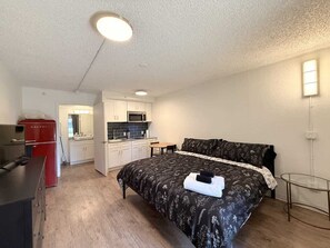 Un dormitorio con cama, una mesita y una kitchenette al fondo.