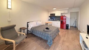 Una stanza compatta con un letto, una sedia, un tavolino, angolo cottura con microonde e frigorifero rosso, e una TV.