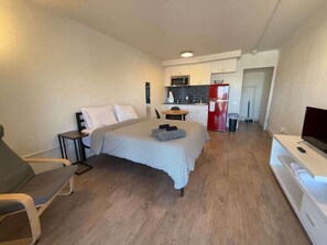 Ein kleines Zimmer mit einem Bett, einem Fernseher, einem kleinen Tisch und einer Küchenzeile im Hintergrund.