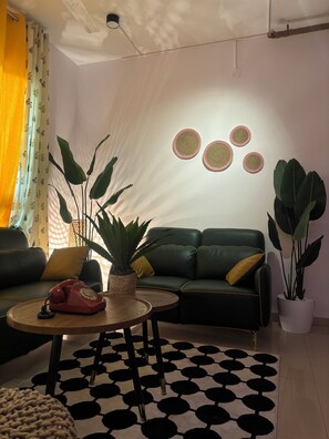 A living room with a green sofa, a round wooden coffee table, a red rotary phone, and large potted plants. - Stylish Boho 2BHK• escape with open creek views•Thane (Thane)