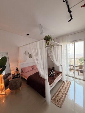 Un dormitorio con cama, mesita de noche, planta colgante, ventana con cortinas y vistas al exterior.