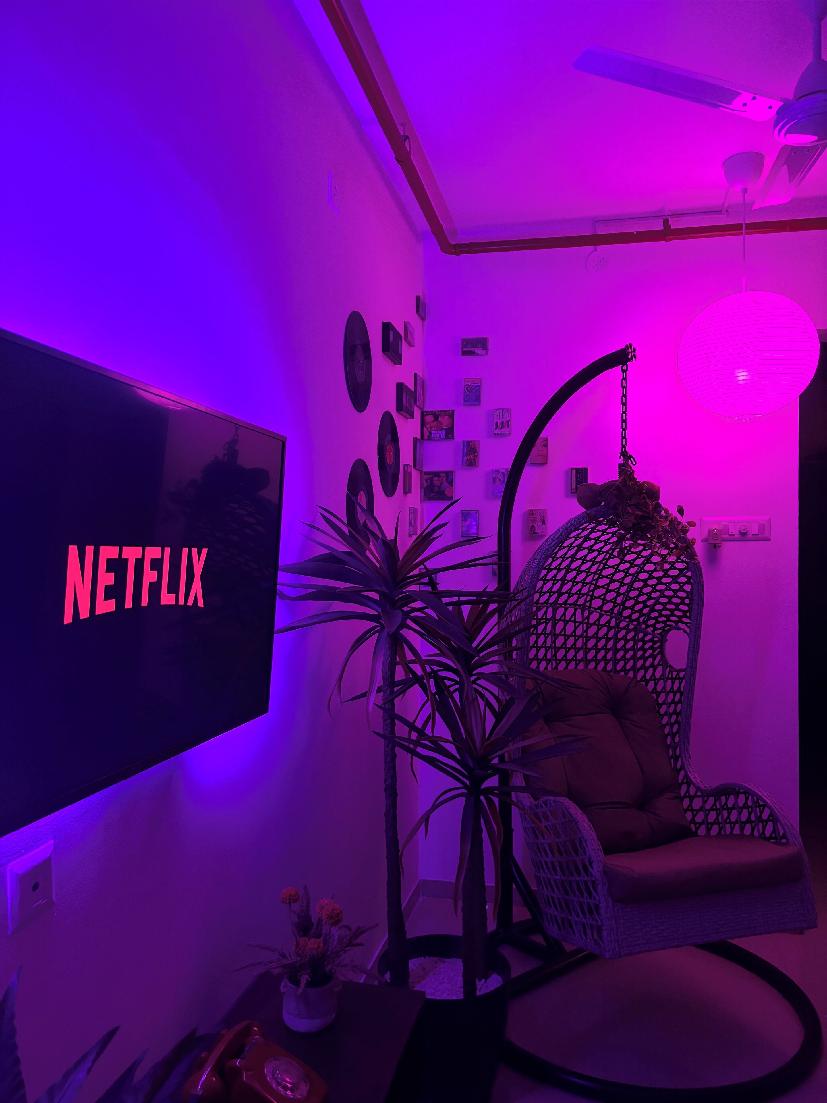 A room with a TV displaying 'NETFLIX,' a ceiling fan, a hanging lamp, a cushioned chair, and wall decorations.