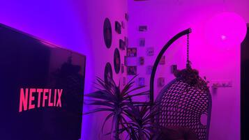 A room with a TV displaying 'NETFLIX,' a ceiling fan, a hanging lamp, a cushioned chair, and wall decorations.