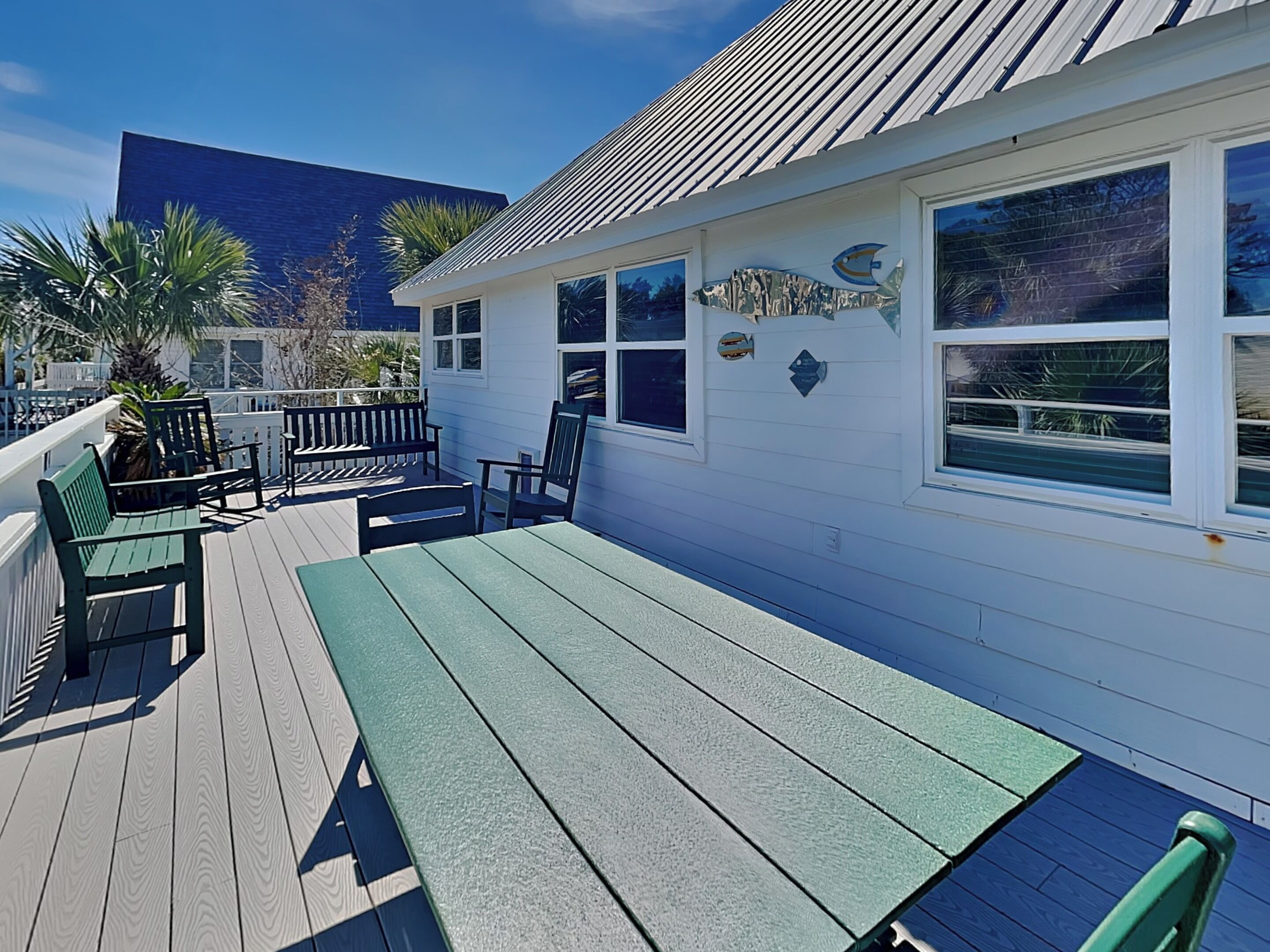 A deck with green benches and a table, a white building with windows, and a blue sky.