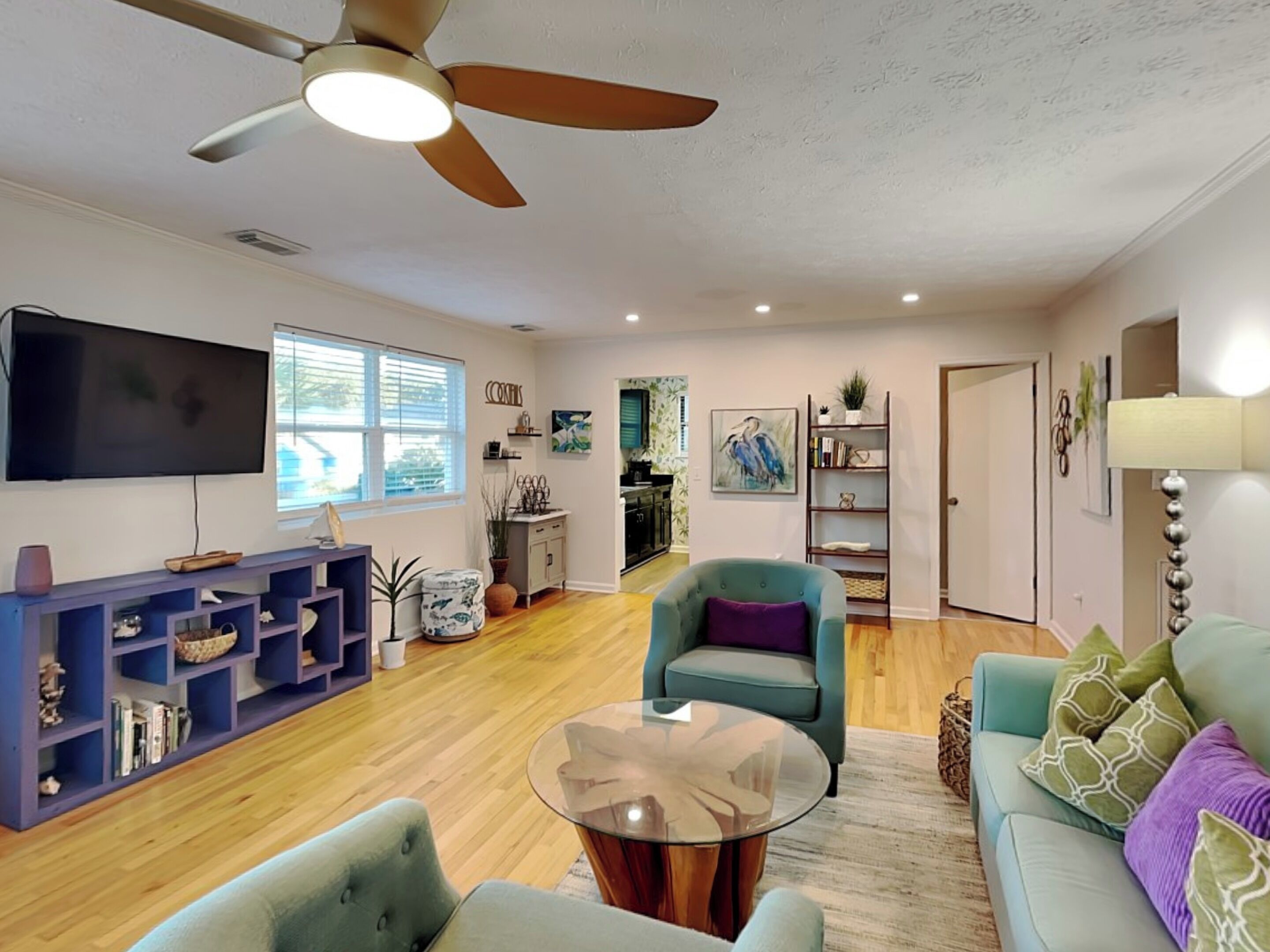 Un salon doté d’un ventilateur de plafond, d’un téléviseur à écran plat, d’un canapé bleu, d’un fauteuil turquoise et d’une table basse en verre.