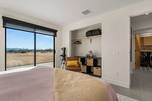 A bedroom with a large bed, a seating area with a chair and cushions, a wardrobe, and a view of the outdoors. - Modern on Mars | MCM remodeled | SPA | Fire-pit! (San Bernardino County)