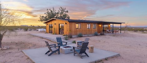 Una cabaña de madera con un porche cubierto, mobiliario exterior y un paisaje desértico al fondo.