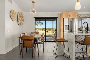 A modern kitchen with a large island, a dining area with a table and chairs, and a view through a glass door. - Modern on Mars | MCM remodeled | SPA | Fire-pit! (San Bernardino County)