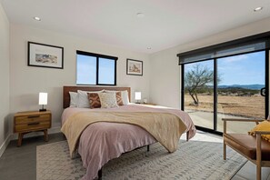 A bedroom with a large bed, a nightstand, a chair, and a view of the outdoors through a sliding door. - Modern on Mars | MCM remodeled | SPA | Fire-pit! (San Bernardino County)