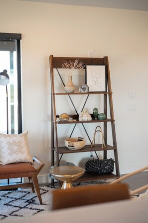 A room with a wooden ladder shelf holding decorative items, a cushioned chair, a small round table, and a patterned rug. - Modern on Mars | MCM remodeled | SPA | Fire-pit! (San Bernardino County)