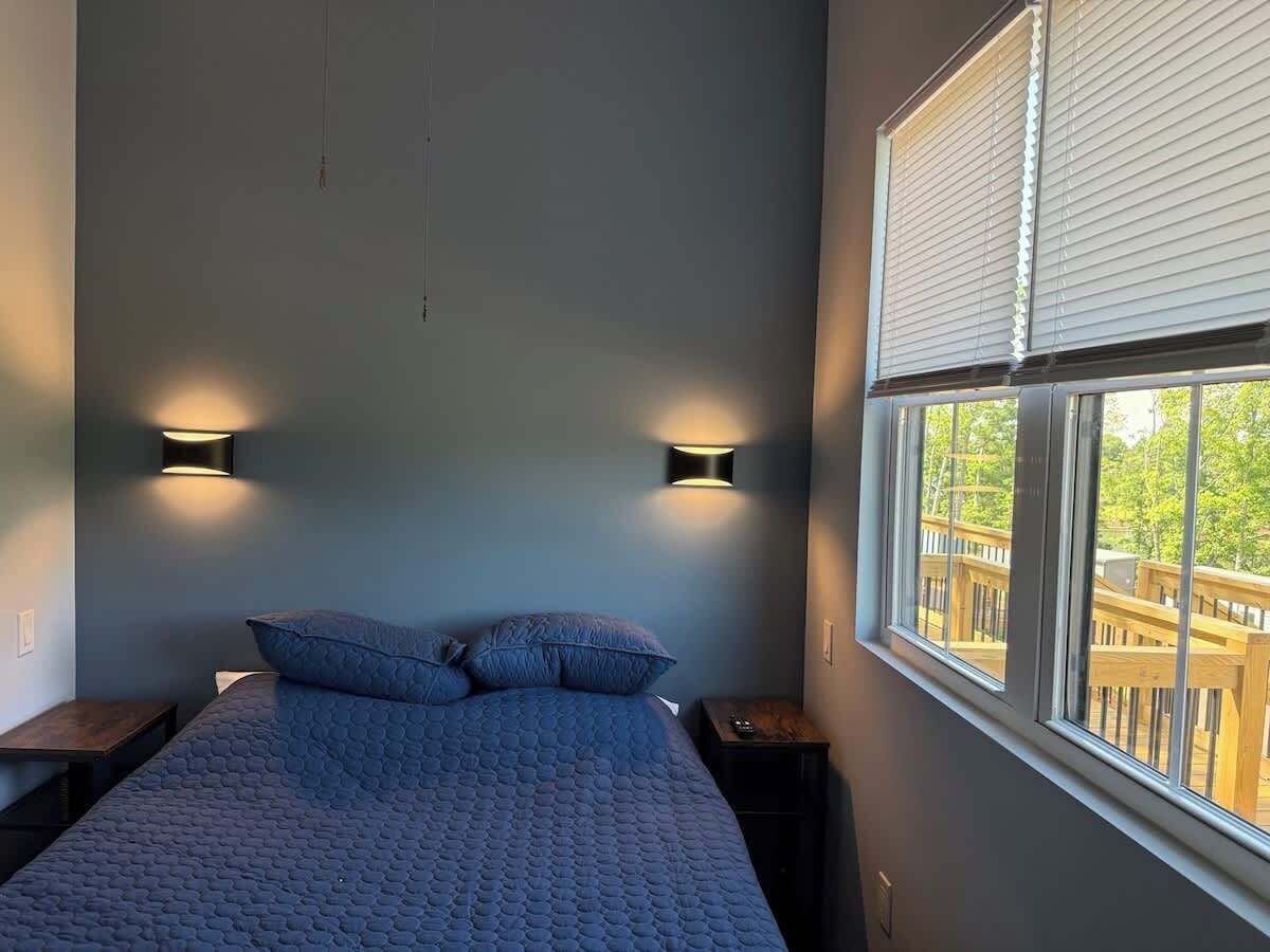 A bedroom with a bed, two wall-mounted lights, a window with blinds, and a view of a deck.