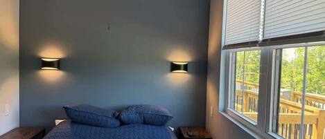 A bedroom with a bed, two wall-mounted lights, a window with blinds, and a view of a deck.