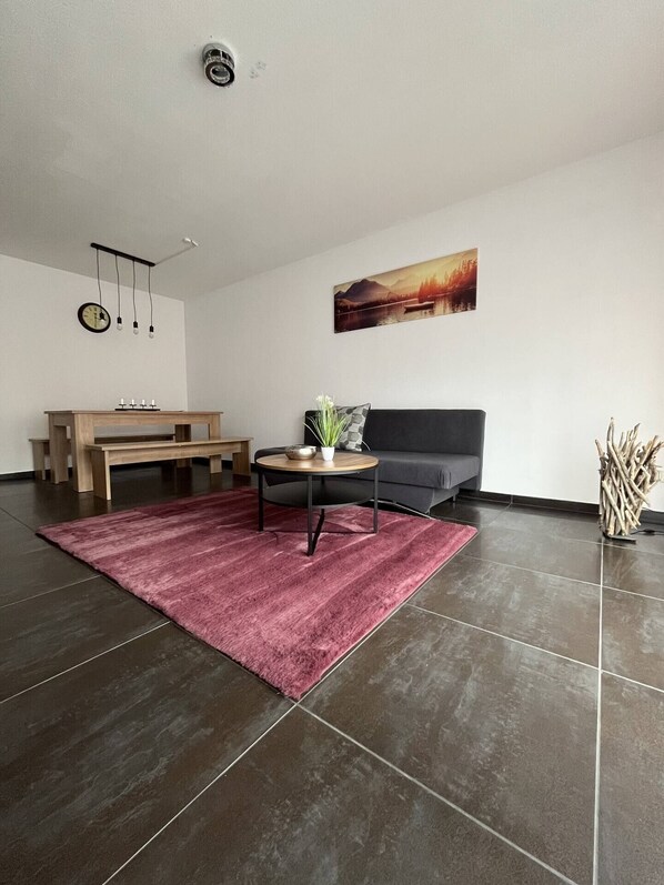 Un salon moderne avec un sol carrelé foncé, un banc en bois, une table ronde ornée d’une plante, un canapé gris et un tableau accroché au mur.