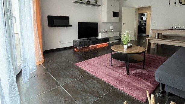 A modern living room with a flat-screen TV, a dining area, and a kitchen in the background. - Modern 3 room apartment | subway fair | parking lot (Fürth)