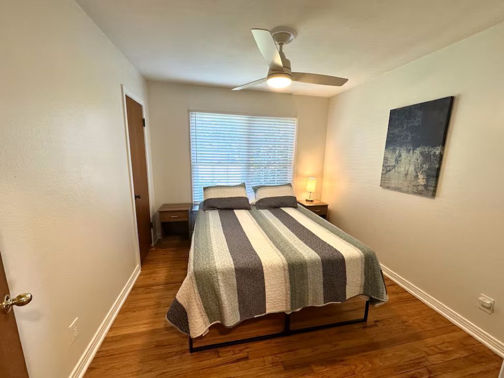 A bedroom with a bed, a ceiling fan, a window with blinds, a nightstand, and a wall-mounted artwork.