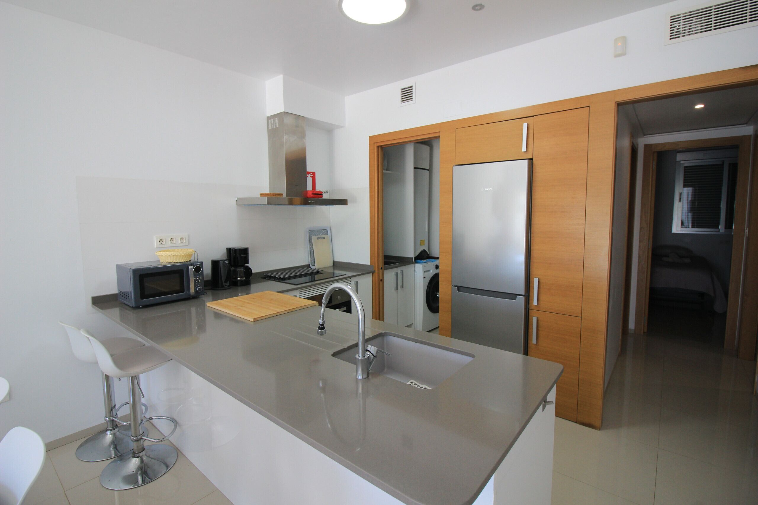 A modern kitchen with a stainless steel sink, a microwave, and a coffee maker. There are wooden cabinets and a wooden door leading to another room.
