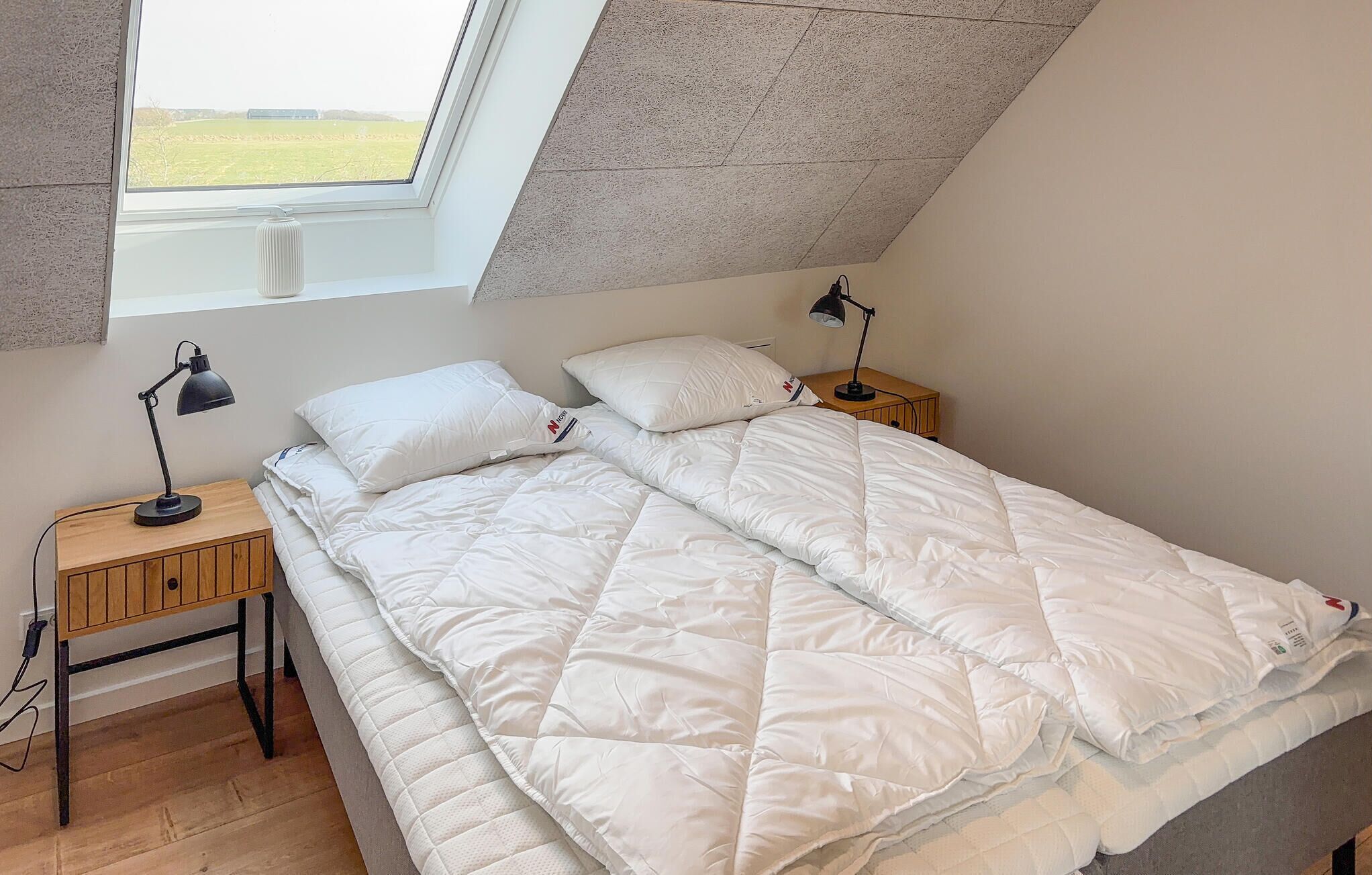 Quarto com teto inclinado, cama com roupa de cama branca, duas luminárias de cabeceira e vista para um campo pela janela.