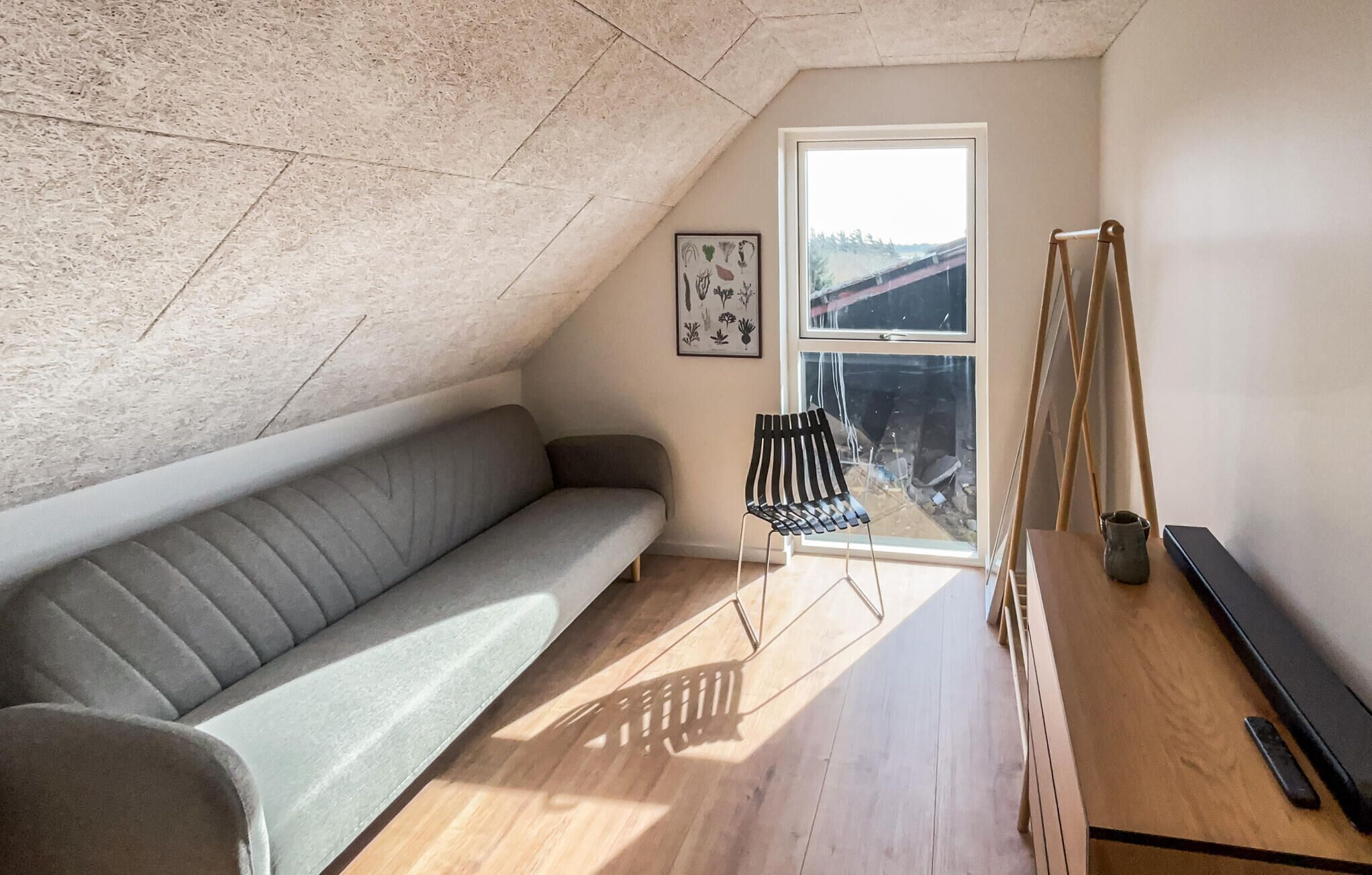 Een loft-achtige kamer met een schuine kap, een grijze bank, een zwarte stoel, een houten tafel en een groot raam.