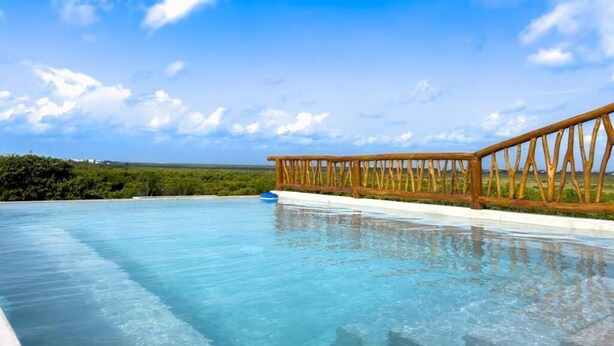 A clear swimming pool with a wooden deck railing, surrounded by greenery and a clear sky. - Casa Daniela  (Puerto Morelos)