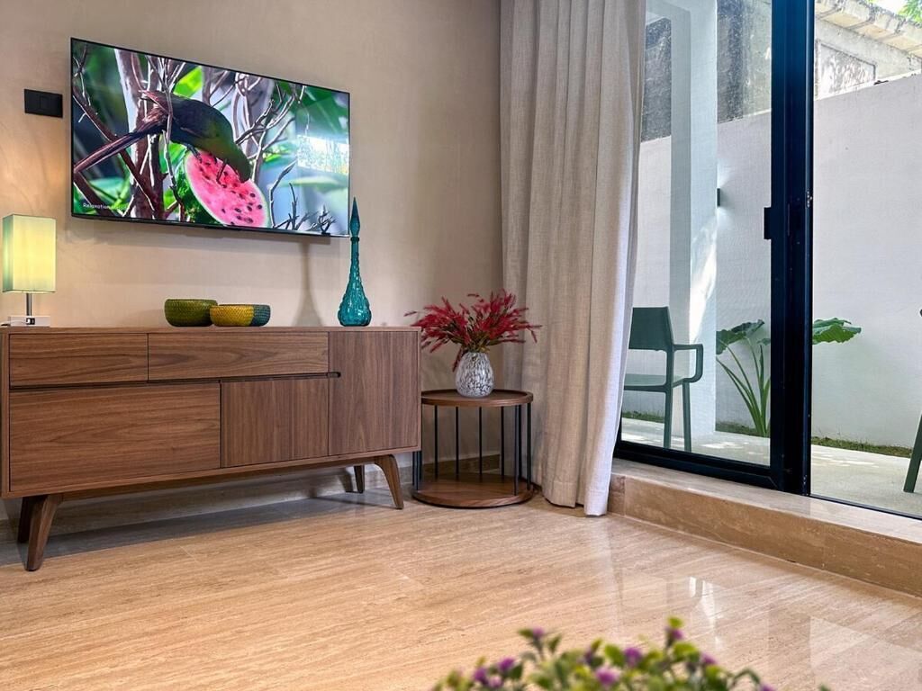 A modern living room with a wooden cabinet, a flat-screen TV, a small round table with a vase, and a view through a glass door.