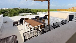 An outdoor patio with a wooden table and chairs, a thatched roof, and a view of greenery and a distant bridge. - Casa Daniela  (Puerto Morelos)