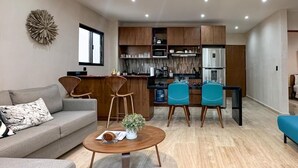 A modern living room with a sofa, coffee table, and chairs, a kitchen area with wooden cabinets and appliances, and a decorative wall piece. - Casa Daniela  (Puerto Morelos)