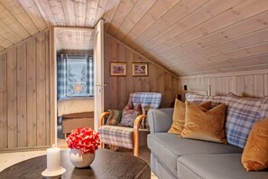 A cozy cabin interior with a loft bed, a sofa, a chair, and a table with a candle and flowers.