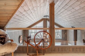 A high-ceilinged room with wooden beams, a large spinning wheel, and a view of snow-covered trees through large windows.