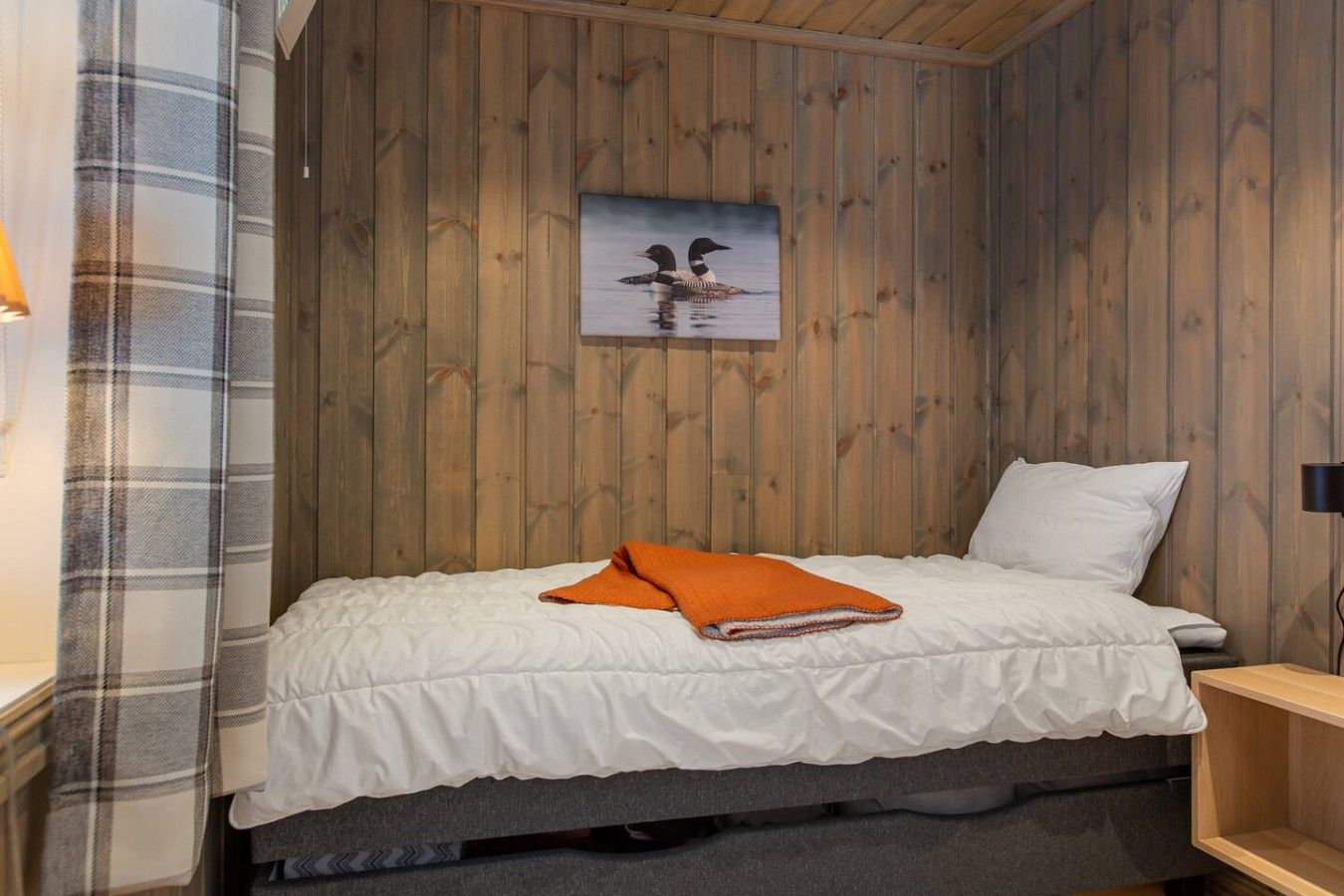 A single bed with a white duvet and an orange blanket, a wooden headboard, and a painting of ducks on the wall.