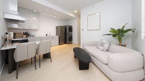 Et moderne køkken med hvide skabe, et spiseområde med et bord og stole, en sofa og en potteplante.