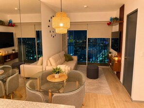 A modern living room with a glass dining table, a sofa, and a hanging light fixture.