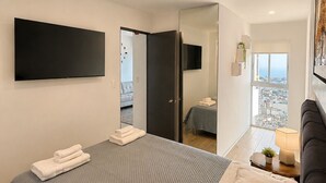 A modern bedroom with a large bed, a flat-screen TV, a door leading to a balcony, and a city view.