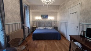 A bedroom with a large bed, a chandelier, a desk, and a chair. - Immerse yourself in this spacious 26m² room inspired by the mastery of light. (Giverny)