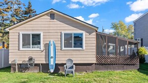 A single-story house with a covered porch, a lawn, and a surfboard leaning against the house. - Beach Cottage w Hot Tub & Screened Porch! (Old Orchard Beach)