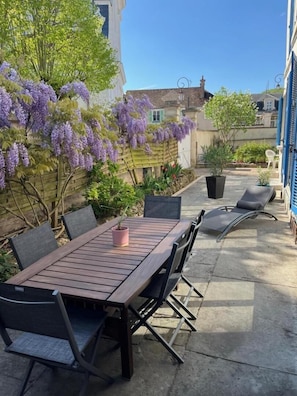 Een terras met een houten tafel en stoelen, omgeven door planten en een bloeiende paarse klimop.