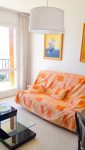 A living room with a sofa, a glass coffee table, a painting, and a window with curtains. - Golfe Juan charmant appartement garage proche mer (Vallauris)