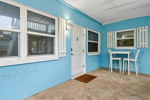 Una habitación con pared azul, una puerta blanca, dos ventanas con contraventanas, una pequeña mesa con sillas y un felpudo marrón.
