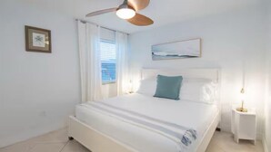 Et soveværelse med en seng, en loftsvifte, et vindue med gardiner, et natbord med en lampe og et billede i ramme på væggen.