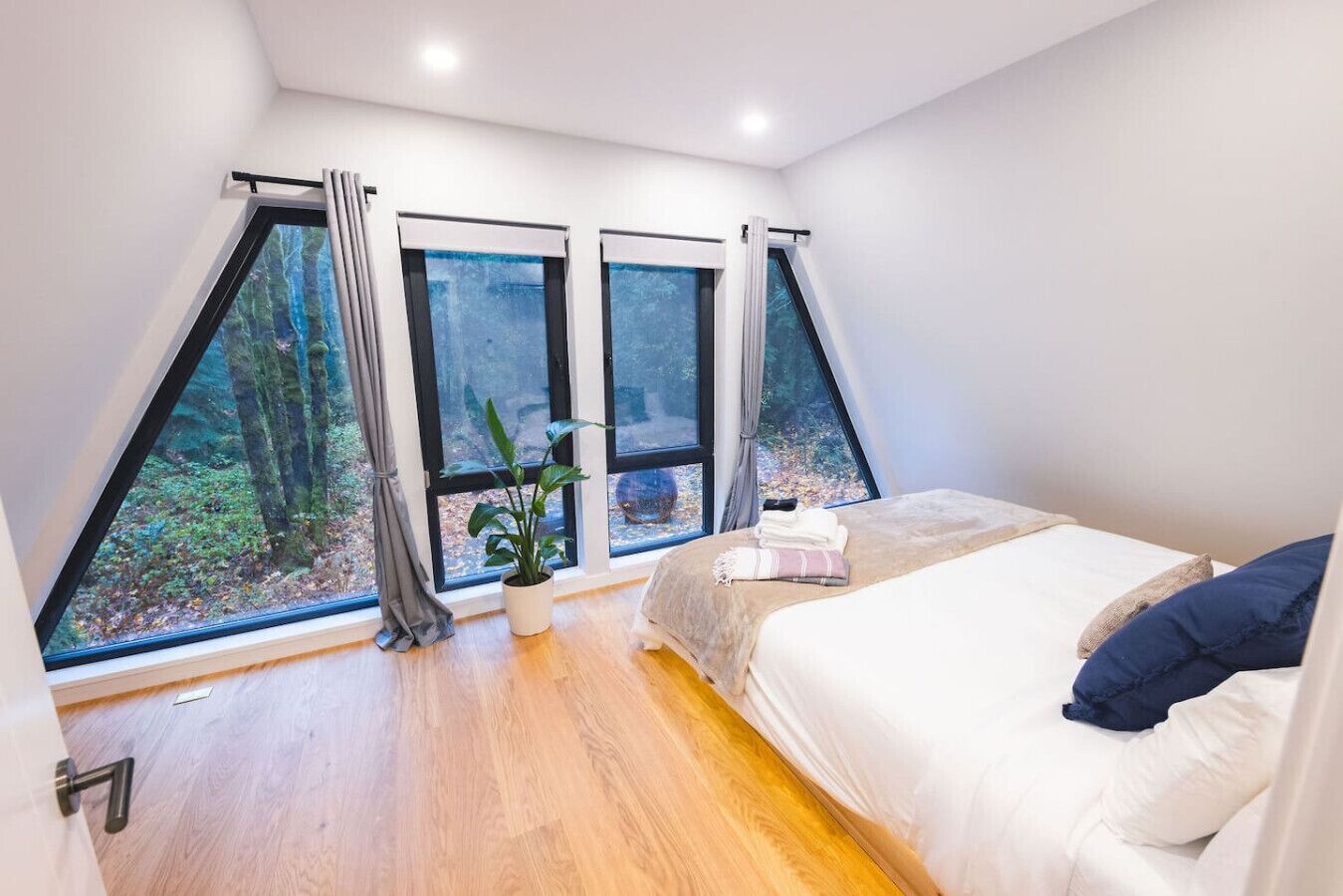 Un dormitorio con una cama grande, una planta en maceta y tres ventanas con vistas a un bosque.
