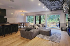 A modern living room with a sectional sofa, a dining area with a bar, and large windows offering a view of the outdoors. - The Adventure Frame- Hot Tub + Sauna + Cold Plunge (Sultan)