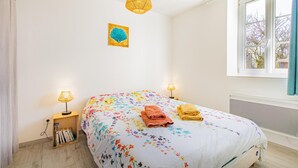 Une chambre à coucher avec une couvre-lit à motifs floraux, deux lampes de chevet, une fenêtre avec des rideaux et une petite table de chevet sur laquelle sont posés des livres.