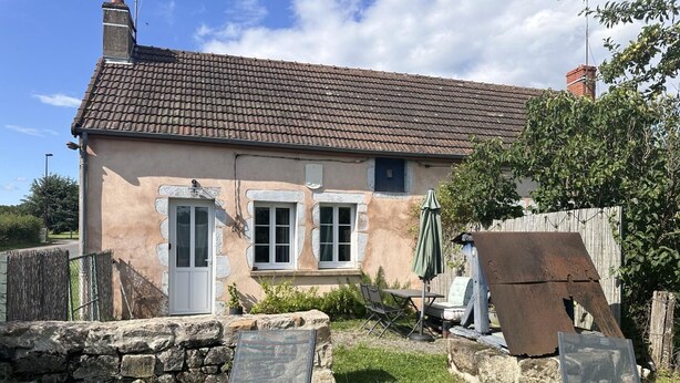 Une maison avec un toit en tuiles, un mur en pierre et un petit jardin agrémenté d’une table et de chaises.