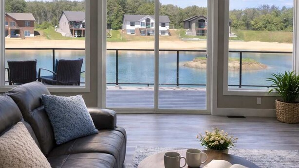 Een ruime kamer met een groot raam dat uitkijkt op een meer en huizen, een leren bank en een ronde salontafel met een boek en twee mokken.
