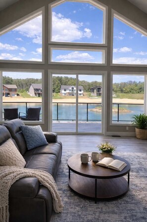 A spacious room with a large window overlooking a lake and houses, a leather sofa, and a round coffee table with a book and two mugs. - Beach House by Castle Rock Lake | Fall Lake Escape + Fire Pit (New Lisbon)