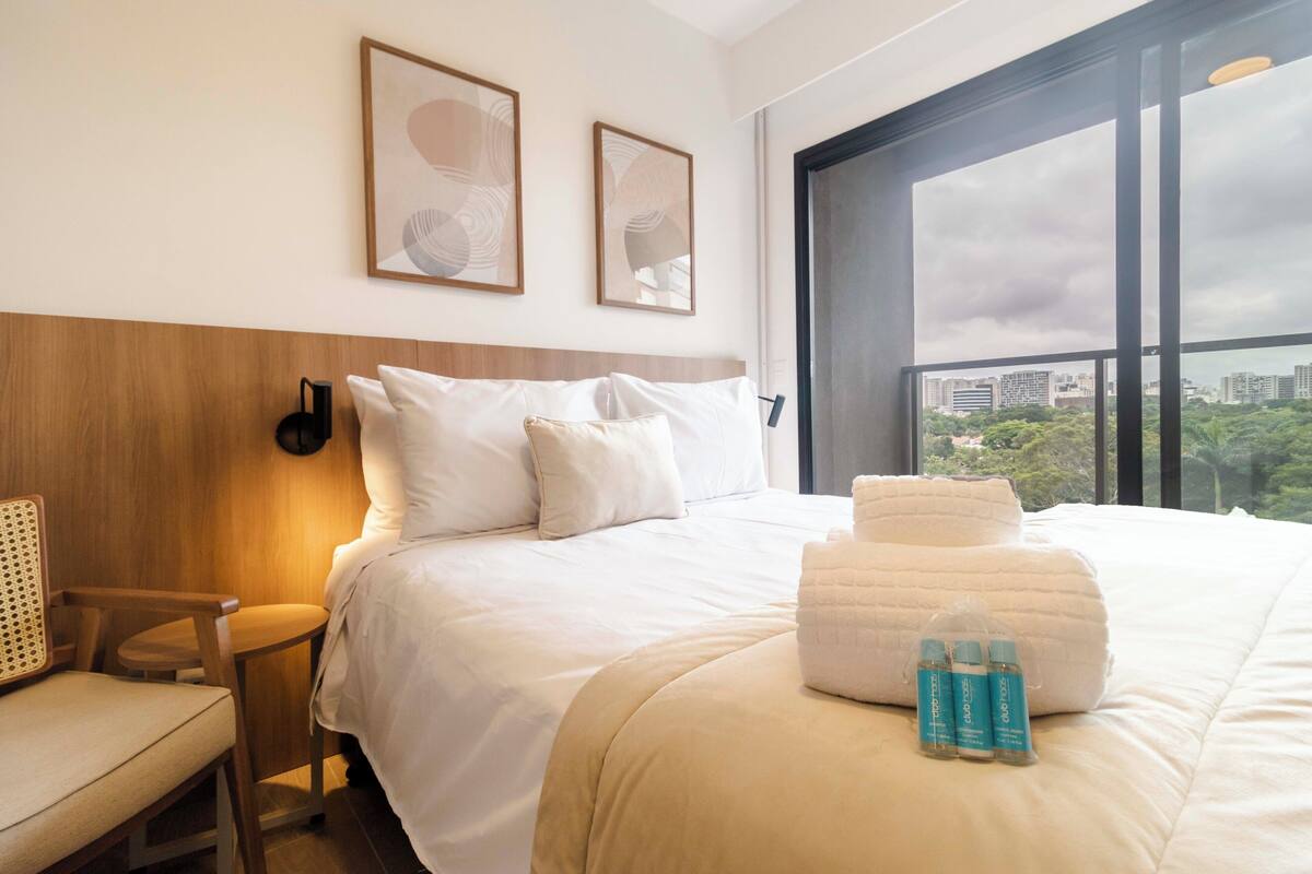 A hotel room with a large bed, a wooden headboard, a window with a city view, and two framed pictures on the wall.