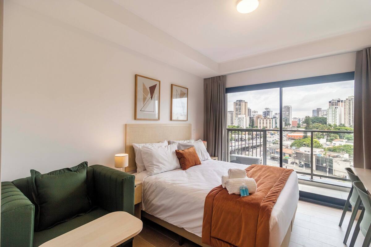 A hotel room with a bed, a green sofa, a dining table, and a large window with a city view.