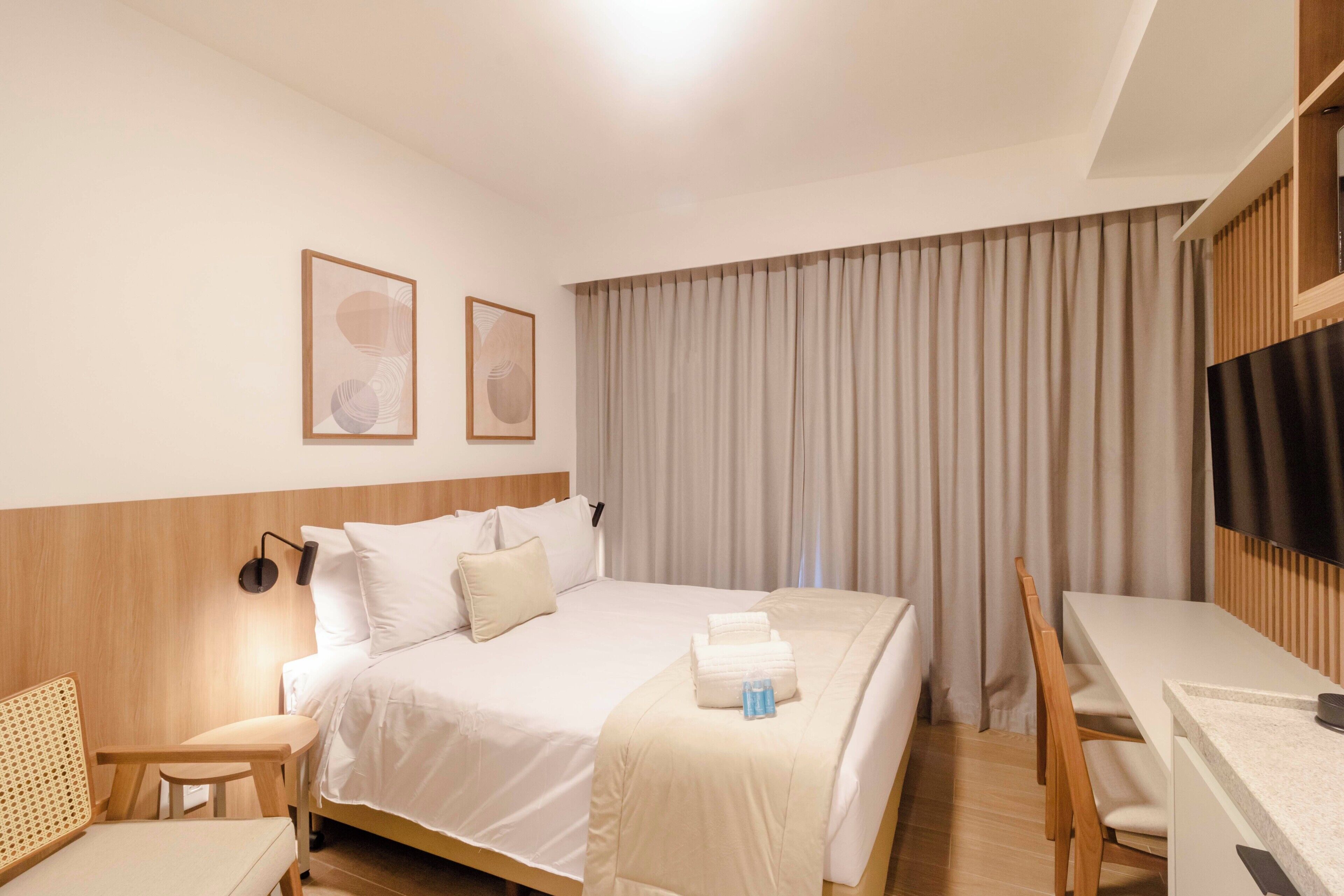 A hotel room with a bed, a desk, a chair, a small table, and a TV.