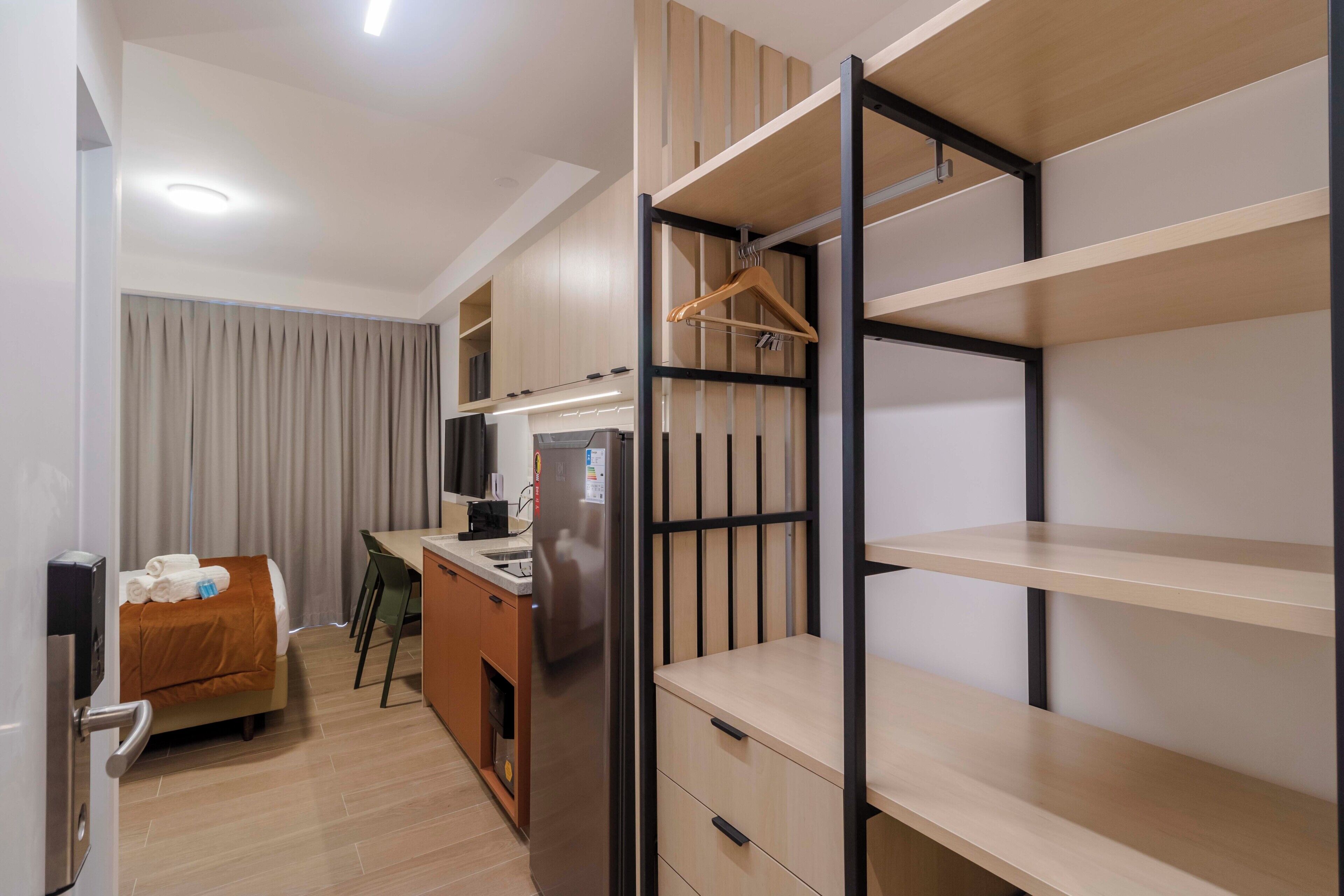 Una habitación de hotel compacta con kitchenette, una cama con manta marrón, perchero y armario empotrado con varias repisas.