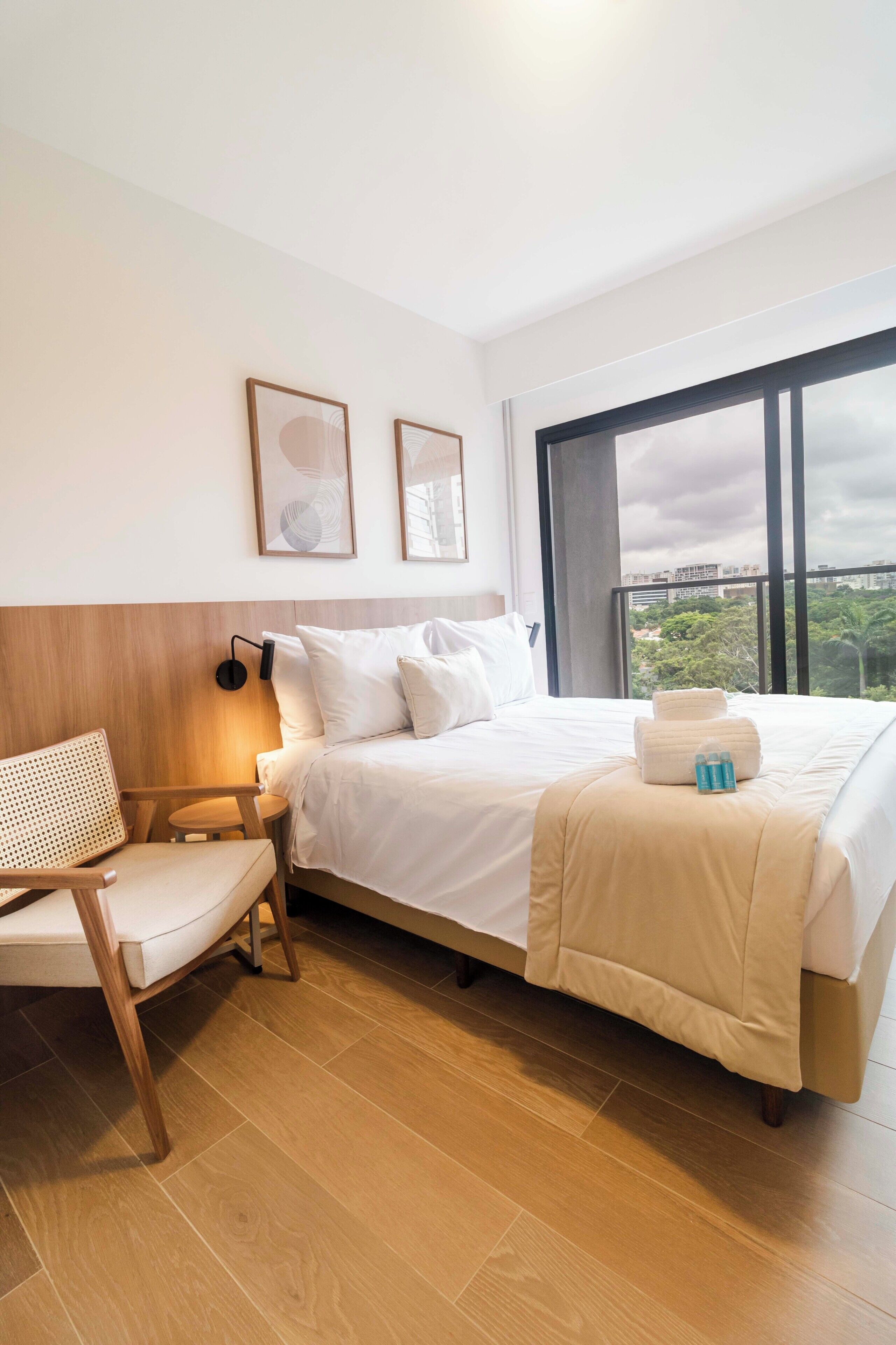 A hotel room with a bed, a chair, a bedside table, and a large window.